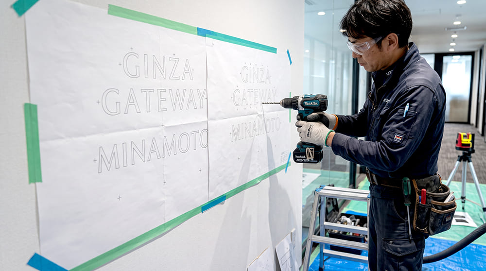 看板職人が原寸原稿シートを壁面に貼りピン立て用ボルトの位置決め穴あけを行う施工風景